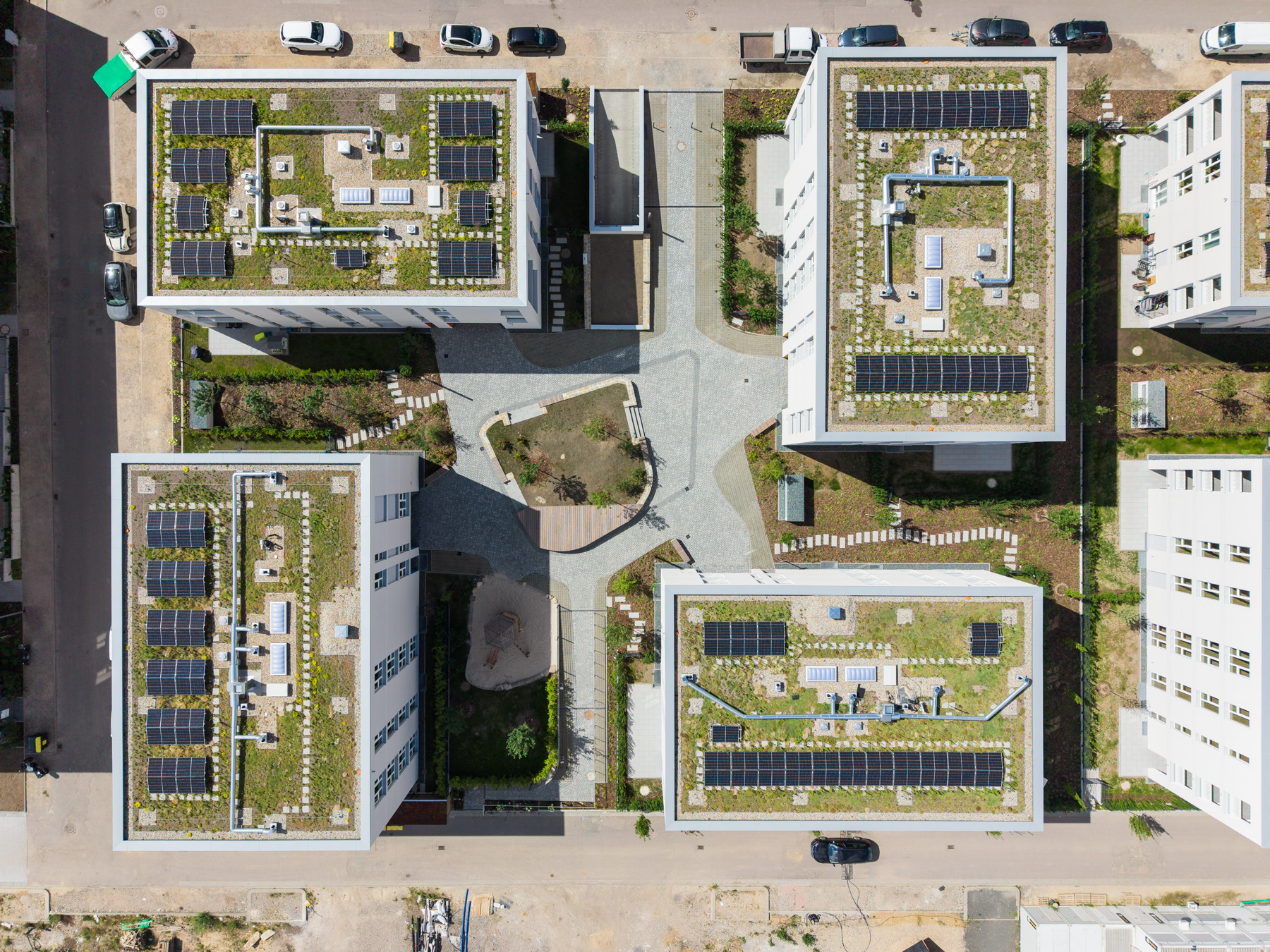 Luftaufnahme von vier weißen Wohngebäuden mit begrünten Flachdächern und Solarmodulen rund um einen gepflasterten Innenhof mit Grünfläche und Sandspielplatz