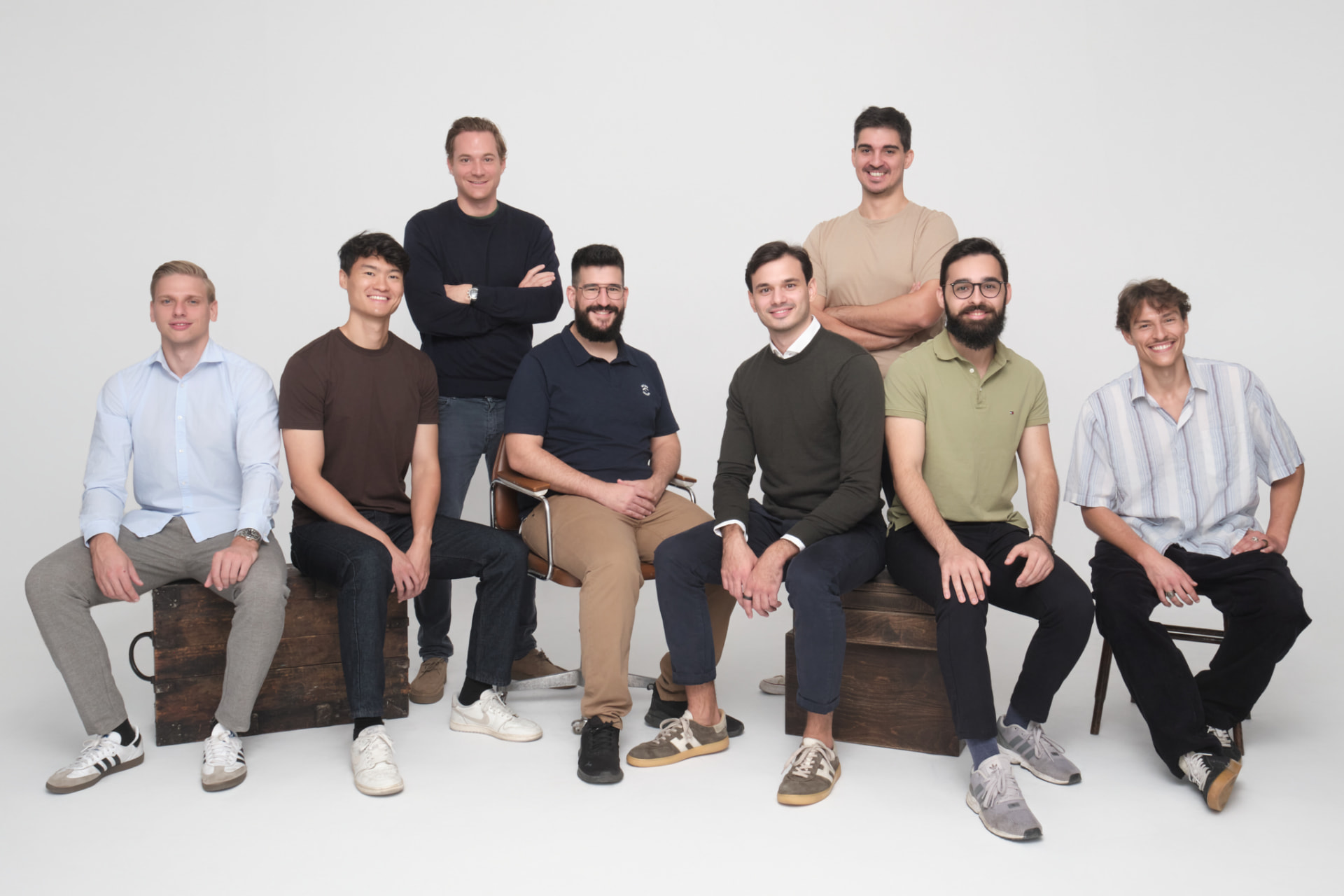 Gruppenbild des Afori-Teams: neun Männer vor hellgrauem Studiohintergrund, sieben sitzen auf Holzkisten und Stühlen, zwei stehen mit verschränkten Armen dahinter