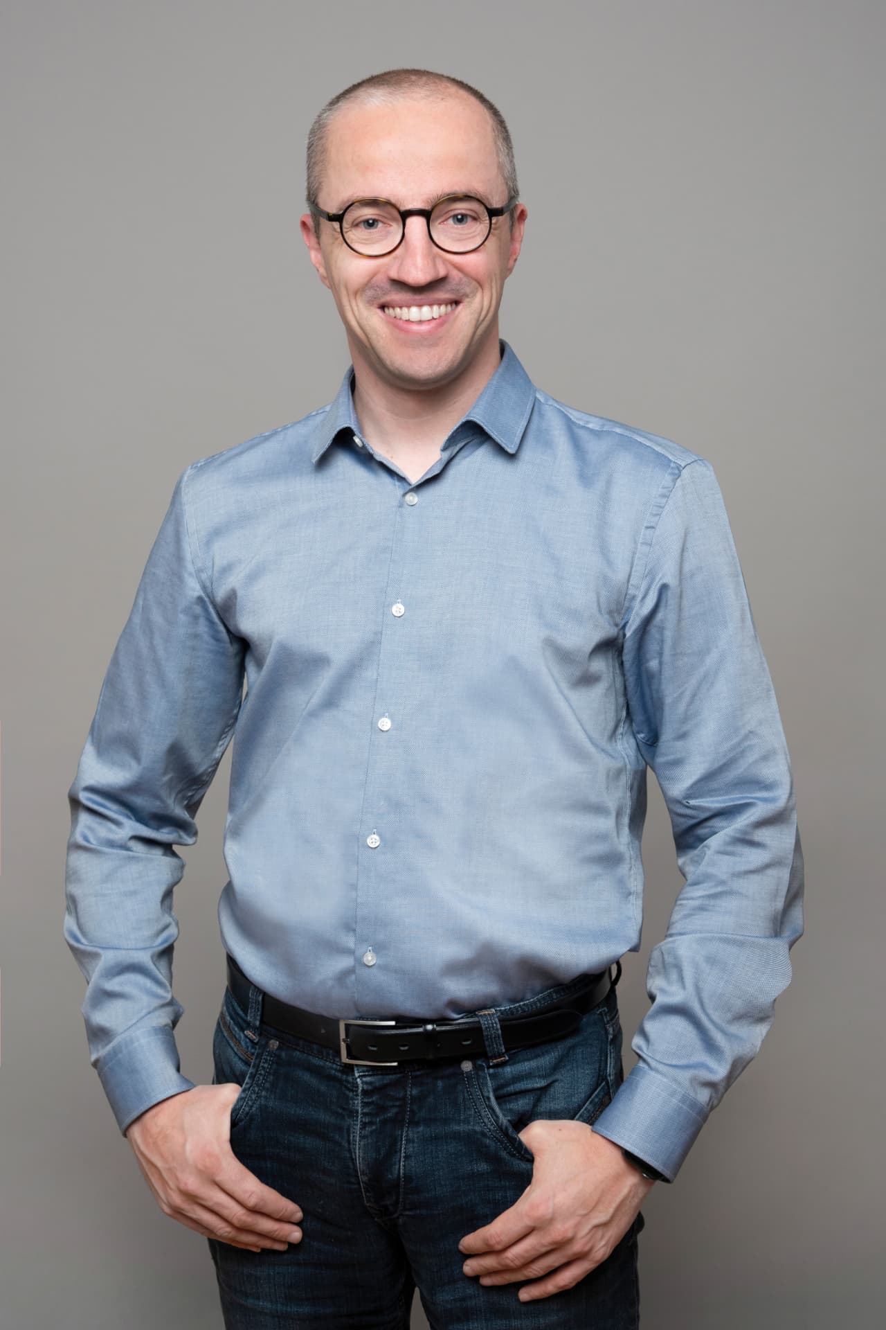Porträt von Stefan Unterthiner, CTO von in-manas: lächelnder Mann mit runder Brille, blauem Hemd und Jeans vor grauem Hintergrund