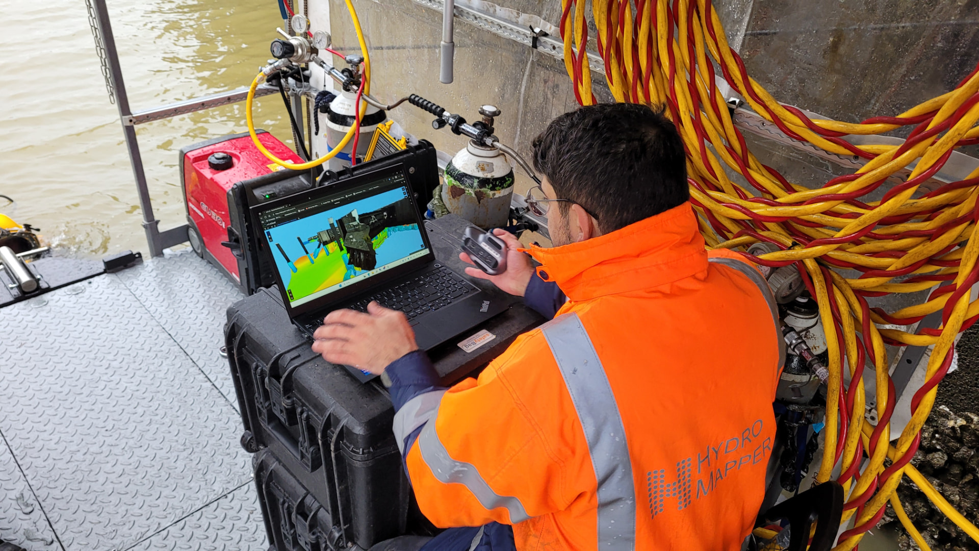 Mitarbeiter in oranger Warnjacke mit HydroMapper-Logo sitzt an Bord eines Messschiffs vor einem Laptop und betrachtet ein farbiges 3D-Modell einer Uferwand; daneben Pressluftflaschen, ein Generator und aufgerollte Versorgungsschläuche.