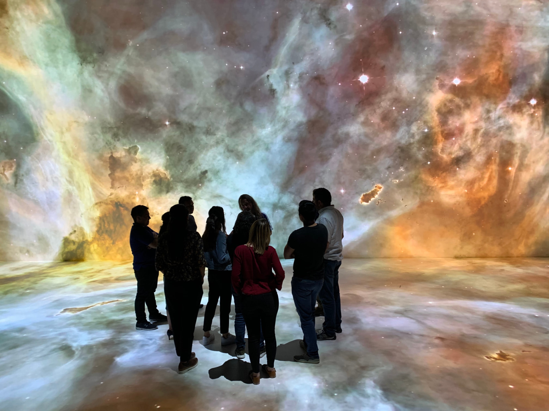 Besuchergruppe steht in einem Deep Space, umgeben von einer großflächigen Projektion eines farbigen Sternennebels auf Wand und Boden.