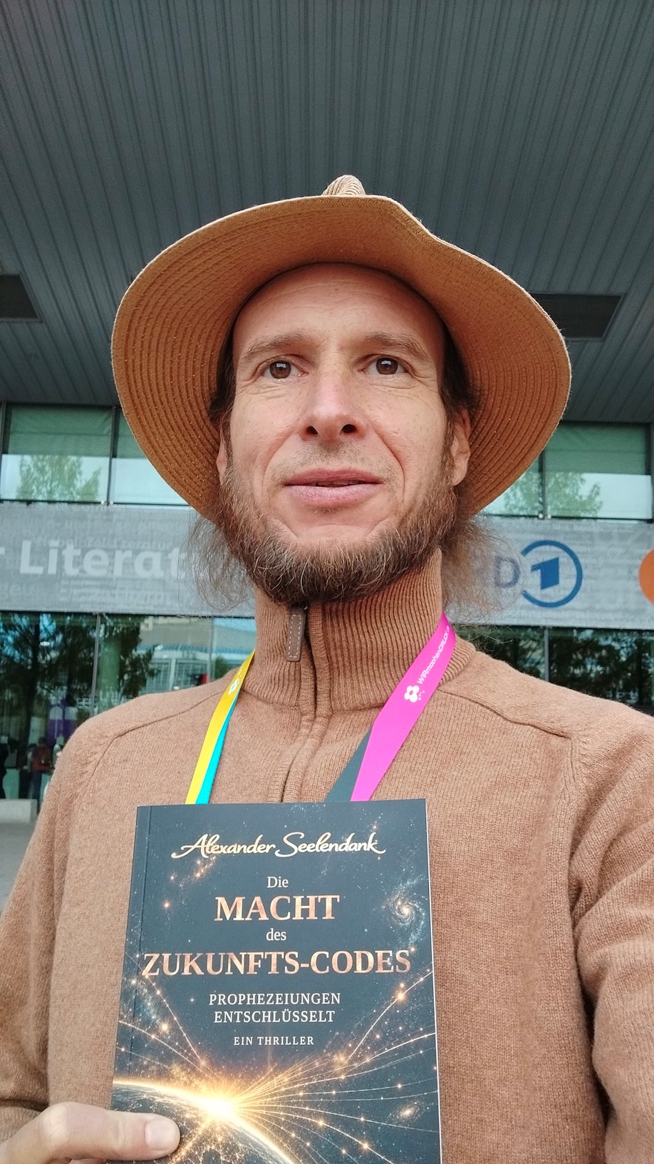 Alexander Seelendank mit Hut und Messe-Umhängeband hält sein Buch „Die Macht des Zukunfts-Codes“ vor einem Hallen-Eingang der Frankfurter Buchmesse